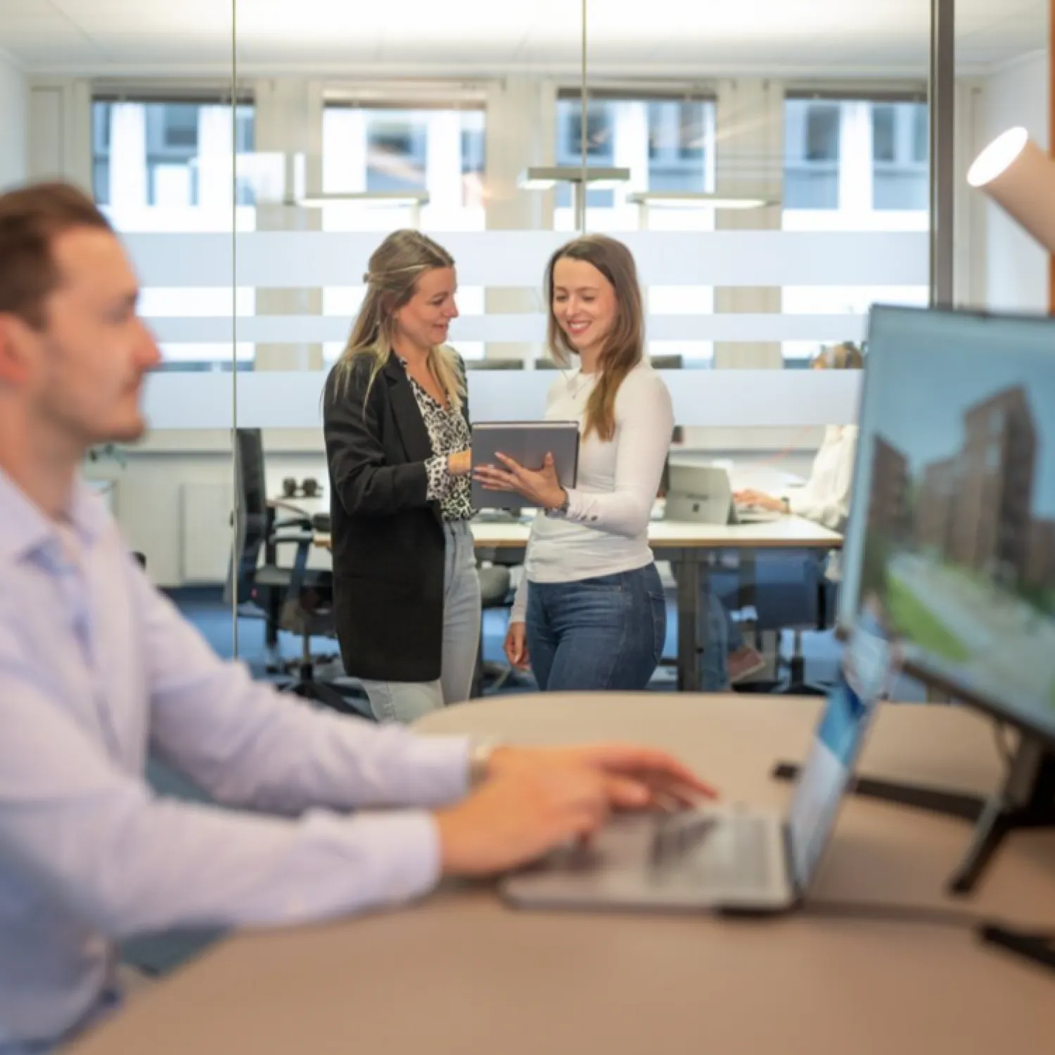 Mann am Schreibtisch mit Computer und im Hintergrund zwei Frauen mit Tablet